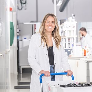 Leende kvinna i vit laboratorierock kör en vagn med plastlådor genom en ljus laboratoriekorridor